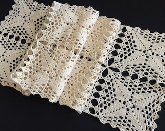 May include: Cream-colored, intricately crocheted table runner. The openwork design features floral and geometric patterns, with a scalloped edge. The runner is folded, showcasing the detailed craftsmanship and texture of the handmade textile.
