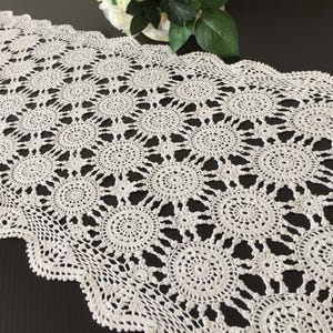 Puede incluir: Un camino de mesa rectangular de ganchillo en color blanco roto con un patrón repetitivo de motivos circulares. El camino presenta un borde festoneado con intrincadas puntadas caladas en toda la tela, creando una textura delicada similar al encaje.