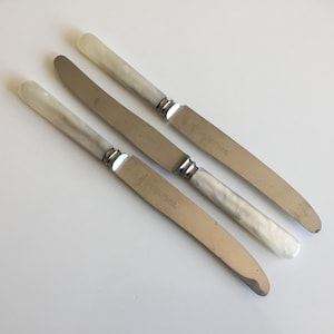 May include: Three vintage stainless steel knives with pearlescent white handles. The blades are silver and the handles have a marbled appearance. The knives are arranged diagonally on a white surface. Each blade has text.