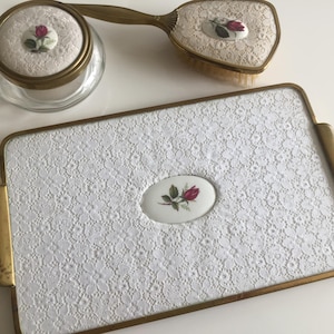 May include: A vintage vanity set comprising a powder jar, brush, and tray. The set features a white lace pattern with brass trim. Each item has an oval porcelain insert with a red rose motif.