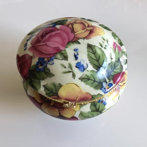 May include: A round porcelain trinket box with a cream-colored base featuring a vibrant floral pattern of pink and yellow roses, green leaves, and small blue flowers. The lid is hinged with a gold-tone metal rim and a small decorative clasp.