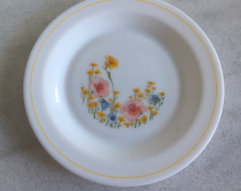 May include: White ceramic plate with a yellow rim and a floral design. The design features pink, yellow, and blue flowers. The plate is round and has a slightly raised edge.