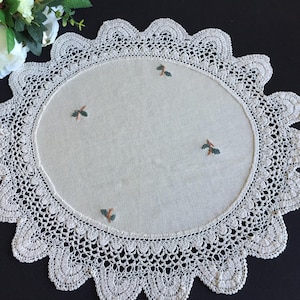 Könnte beinhalten: Ein rundes, cremeweißes Stoffdeckchen mit einem breiten, aufwendig gehäkelten Spitzenrand mit gewelltem Abschluss. Die Mitte des Deckchens besteht aus schlichtem Stoff und ist mit kleinen, verstreuten Stickmotiven aus grünen Blättern und braunen Stielen verziert.