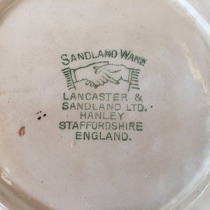 May include: Close-up of a vintage ceramic plate with a cream-colored surface. The plate's underside features the green text "SANDLAND WARE" and "LANCASTER & SANDLAND LTD. HANLEY STAFFORDSHIRE ENGLAND."