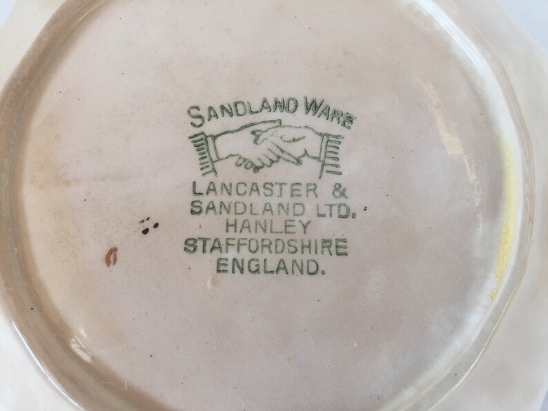 May include: Close-up of a vintage ceramic plate with a cream-colored surface. The plate's underside features the green text "SANDLAND WARE" and "LANCASTER & SANDLAND LTD. HANLEY STAFFORDSHIRE ENGLAND."
