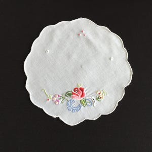May include: A white, scalloped-edge doily with floral embroidery. The embroidery features a red rose, blue swirls, and small flowers in pink, yellow, and green. Three small pink beads are embroidered near the top edge.