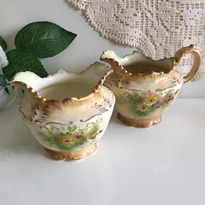 May include: Two vintage cream jugs with a scalloped edge and floral pattern. The jugs are cream-coloured with gold trim and a painted floral design in yellow, pink, and green. One jug has a handle.