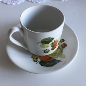 以下が含まれることがあります： 白いセラミックティーカップとソーサーのセット。カップとソーサーは、緑の葉、オレンジと赤のベリーを使ったカラフルなフルーツデザインが特徴です。カップには湾曲したハンドルが付いています。セットは白い表面にあります。