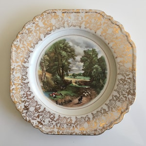 May include: A vintage decorative plate with a landscape scene in the center. The plate has a white background with a gold floral pattern around the rim. The landscape features trees, a river, and figures.