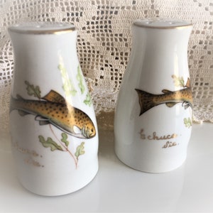 Op de afbeelding: Een paar witte keramische zout- en peperstrooiers met een gouden rand. Elke strooier heeft een handgeschilderd beeld van een forel met bruine en gouden markeringen. De tekst "la chueca" staat op de bodem van de strooiers.