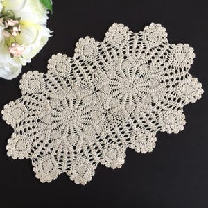 Pode incluir: Toalha de mesa ovalada de crochê creme com borda ondulada. O intrincado design vazado apresenta padrões florais e em forma de coração. A toalha de mesa é exibida em uma superfície preta, com flores brancas no canto superior esquerdo.