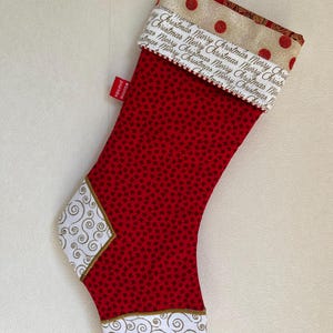 Puede incluir: Un calcetín navideño con cuerpo de tela roja y un pequeño patrón de estrellas negras. El puño es color crema con lunares rojos y el texto Merry Christmas repetido. El talón y la puntera son blancos con espirales doradas y una etiqueta roja.
