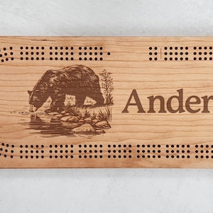 May include: A wooden cribbage board featuring a bear illustration and the name "Anderson" engraved in brown. The board has a series of small holes for peg placement, and a curved oval design.