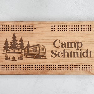 May include: A wooden cribbage board with a camping theme. The board is engraved with a camper, trees, and a campfire. The text "Camp Schmidt" is also engraved on the board.