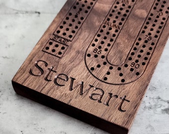May include: A personalized wooden cribbage board with the name "Stewart" engraved on the bottom. The board features a traditional U-shaped track with numbered holes and a "Skunk" area.