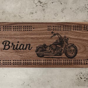 Peut inclure: Un plateau de cribbage rectangulaire en bois avec une finition au grain brun foncé. Le plateau est gravé du prénom Brian en écriture cursive et d'une illustration détaillée de moto, entouré d'une piste continue de trous pour les fiches.