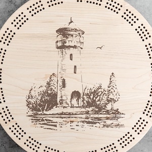 May include: A round wooden cribbage board with a detailed engraving of a lighthouse scene. The board features a series of drilled holes around the perimeter for scoring. The lighthouse is brown, and the board is light-colored.