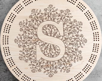 May include: A round wooden cribbage board with a floral design surrounding the letter "S". The board has evenly spaced holes around the perimeter for pegging and is made of light-colored wood.