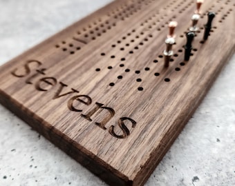 May include: A wooden game board with the name "Stevens" engraved on it. The board has a grid of holes and is used for a game of cribbage. There are several pegs in the holes, some are black, some are silver, and one is rose gold.