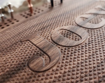 May include: Close-up of a wooden cribbage board with the number "50" carved into the surface. The board has a textured pattern and includes holes for pegs. Various colored pegs are visible.