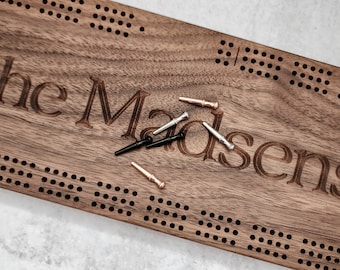 May include: A wooden game board with the name "The Madsens" engraved on it. The board has a grid of holes and comes with several metal and wooden pegs.