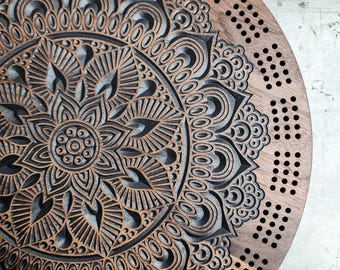 May include: A circular wooden cribbage board with a carved mandala pattern. The board is finished in dark brown and features an elaborate, symmetrical design with drilled holes for pegs. The mandala is the central focus, with detailed geometric patterns.