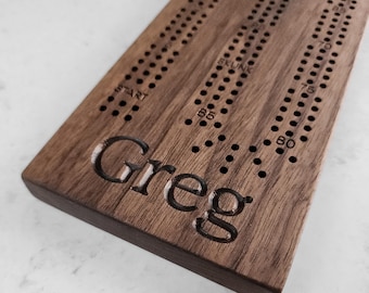 May include: A personalized wooden cribbage board with the name "Greg" engraved on the surface. The board has a dark brown walnut finish and features a traditional cribbage board design with numbered holes and a scoring track.