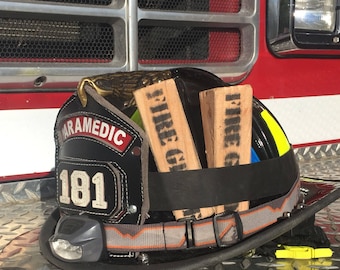 May include: A black firefighter helmet with a gold badge that says "PARAMEDIC 181". The helmet is sitting on a metal surface with a diamond pattern. There are two wooden blocks with the word "FIRE" written on them.