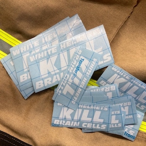 May include: A collection of blue and white transfer stickers with the words "WHITE HELMETS KILL BRAIN CELLS" printed on them. The stickers are on a tan fabric background with yellow and reflective stripes.