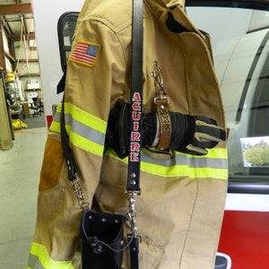 May include: A tan firefighter jacket with yellow reflective stripes and an American flag patch. The jacket has a black strap with a silver buckle and the name "AGUIRRE" printed in red. The jacket is hanging on a white fire truck.