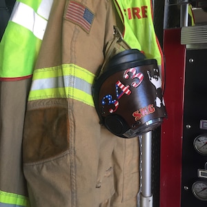 May include: A brown leather firefighter helmet with the number "343" and the letters "SKG" written on it. The helmet is hanging on a hook next to a yellow and brown firefighter jacket with an American flag patch.