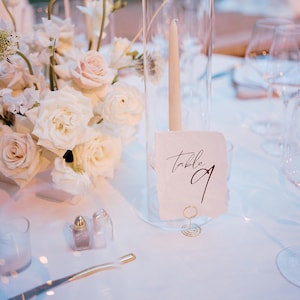 Peut inclure: Une carte de numéro de table blanche avec le texte "Table 9" en calligraphie noire. La carte est maintenue par un clip en métal doré et repose sur une nappe blanche. Un vase en verre haut avec une bougie blanche et des fleurs blanches est en arrière-plan.