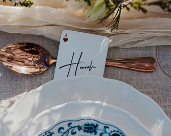 Puede incluir: Una tarjeta blanca con el As de Corazones y el nombre "Hannah" escrito en caligrafía negra. La tarjeta está colocada sobre un plato blanco con un patrón floral azul y blanco. Una cuchara de cobre está colocada junto al plato.