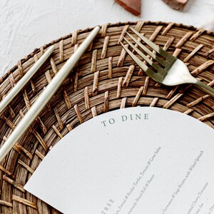 May include: A white paper menu with the text "TO DINE" printed on it. The menu is placed on a woven brown placemat with a gold fork and two silver utensils.
