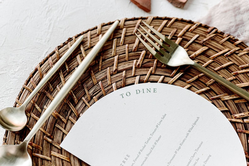 May include: A white paper menu with the text "TO DINE" printed on it. The menu is placed on a woven brown placemat with a gold fork and two silver utensils.