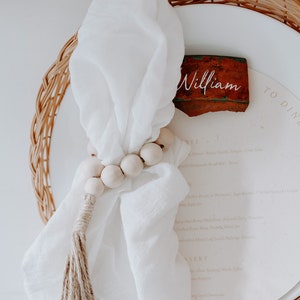 May include: A white linen napkin with a wooden bead garland and a tassel. The napkin is placed on a white plate with a brown clay place card that says "William".