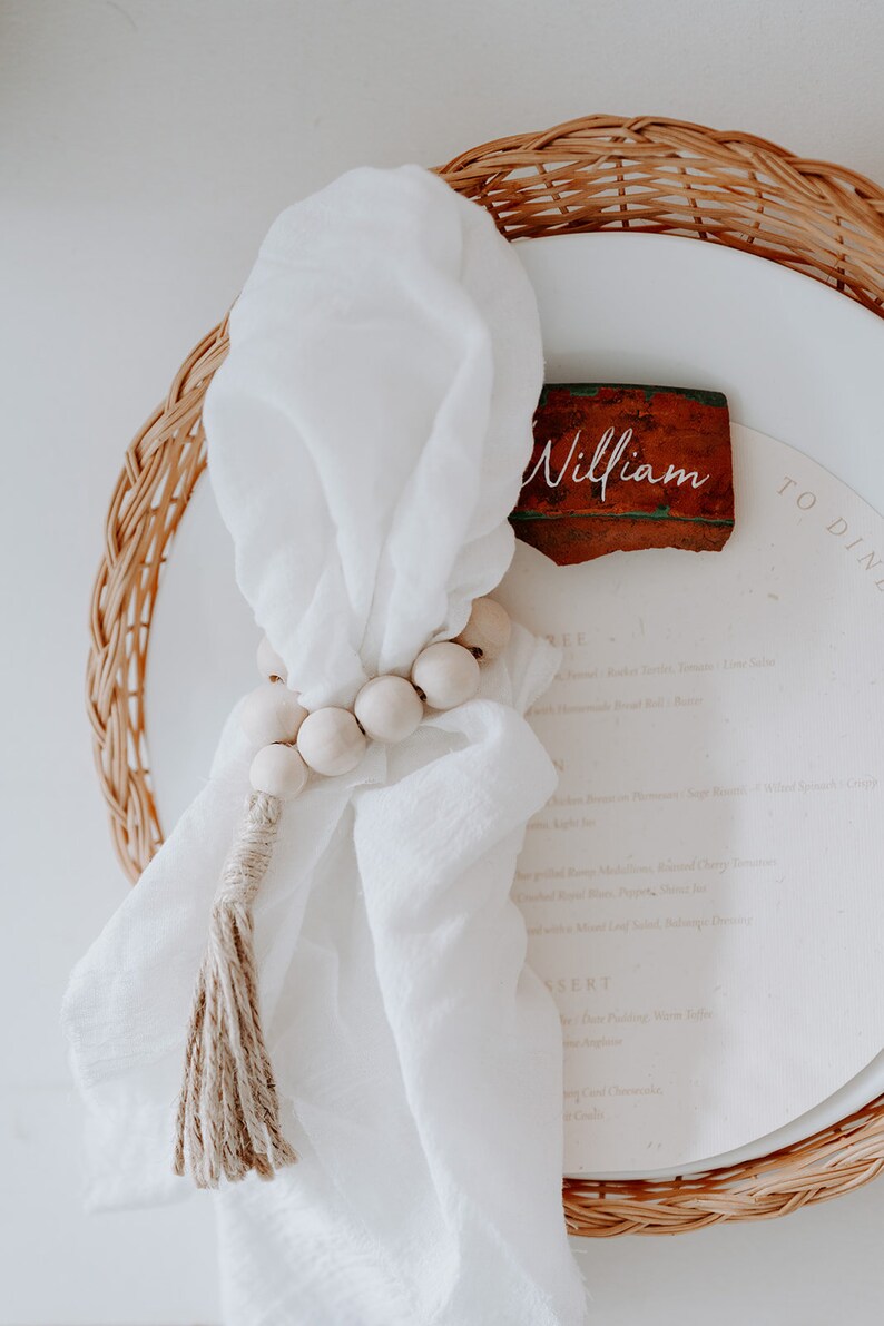May include: A white linen napkin with a wooden bead garland and a tassel. The napkin is placed on a white plate with a brown clay place card that says "William".