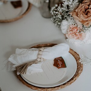 May include: A white napkin with a wooden bead ring and a brown tassel is placed on a white plate with a brown wicker charger. A wooden name card with the name "Courtney" is placed on the plate.