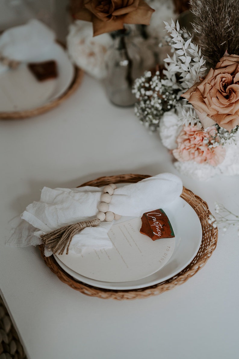May include: A white napkin with a wooden bead ring and a brown tassel is placed on a white plate with a brown wicker charger. A wooden name card with the name "Courtney" is placed on the plate.