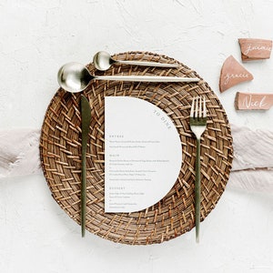 May include: A place setting with a woven placemat, gold cutlery, and a white menu card with the text "To Dine". The place setting is on a white surface with three terracotta place cards with the names "Jamie", "Grace", and "Nick" written on them.