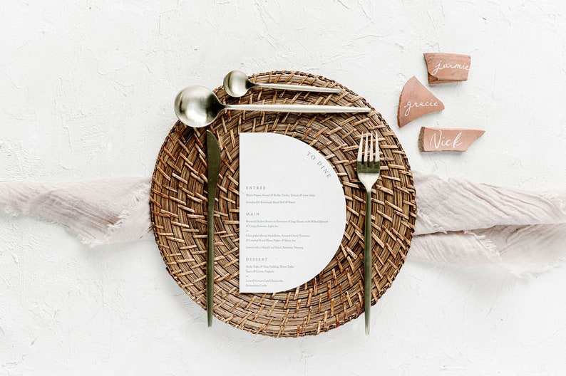 May include: A place setting with a woven placemat, gold cutlery, and a white menu card with the text "To Dine". The place setting is on a white surface with three terracotta place cards with the names "Jamie", "Grace", and "Nick" written on them.
