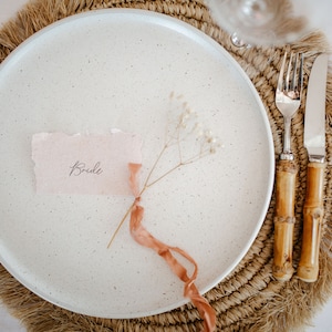 Puede incluir: Un plato blanco moteado con una tarjeta de lugar de papel rosa que dice "Bride" y una ramita de flores blancas secas atada con una cinta rosa. El plato está sobre un mantel individual tejido marrón con flecos.