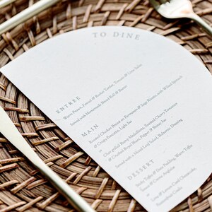 May include: A white menu card with the words "To Dine" at the top. The menu lists the entree, main course, and dessert options. The card is placed on a woven placemat with gold silverware.