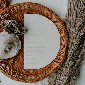 May include: A white paper menu with the text "To Dine" printed on it. The menu is placed on a woven wicker plate with a white flower and green foliage on the left side.