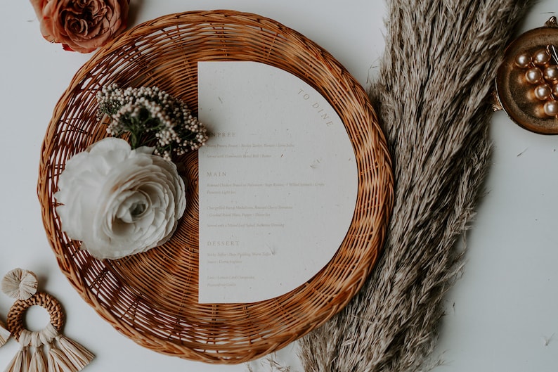 May include: A white paper menu with the text "To Dine" printed on it. The menu is placed on a woven wicker plate with a white flower and green foliage on the left side.