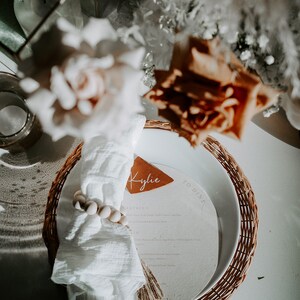 May include: A white napkin with a wooden bead ring sits on a woven placemat with a brown leather name card that says "Kylie" and a white card with the text "To Dinner".