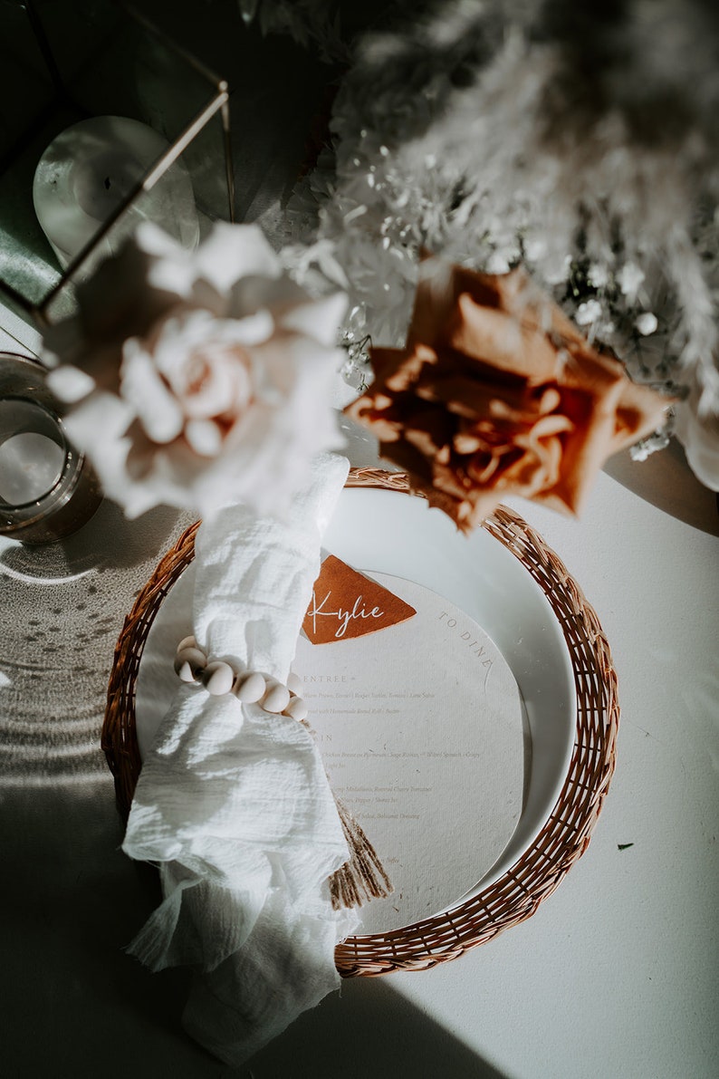May include: A white napkin with a wooden bead ring sits on a woven placemat with a brown leather name card that says "Kylie" and a white card with the text "To Dinner".