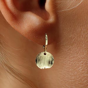 Può includere: Primo piano di un orecchino color oro. L'orecchino è un piccolo disco circolare con una superficie strutturata, appeso a un piccolo cerchio. L'orecchino è indossato su un orecchio, con capelli biondi visibili.