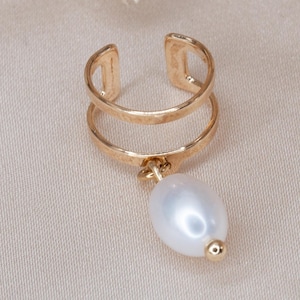 May include: A gold-colored ear cuff with a double-band design. A white, teardrop-shaped pearl dangles from the bottom band. The cuff is set against a neutral background, with dried flowers in the upper corners.