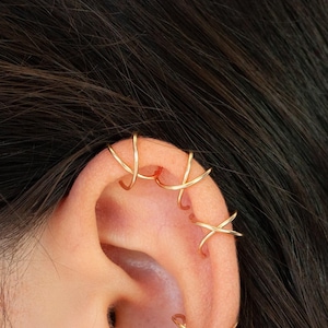 Könnte beinhalten: Nahaufnahme eines Ohrs, das mit mehreren goldenen Ohrringen geschmückt ist. Das Ohr hat kleine Creolen und mehrere Ohrklemmen mit einem X-förmigen Design. Der Schmuck ist vor dunklem Haar und Haut platziert.